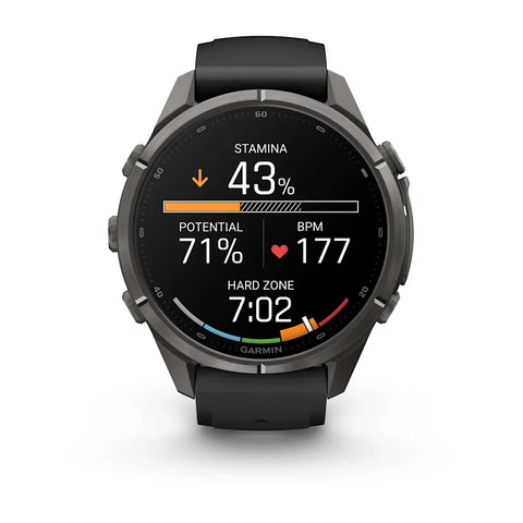 A Garmin smartwatch with a black sport band and a dark gray metallic case, shown against a plain white background. The watch face is active with various fitness metrics displayed.