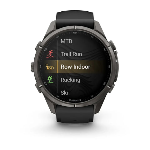 A Garmin smartwatch with a dark gray metallic case and a black strap, shown on a clean white background. The watch screen displays fitness activity options including MTB, Trail Run, Row Indoor, Rucking, and Ski.