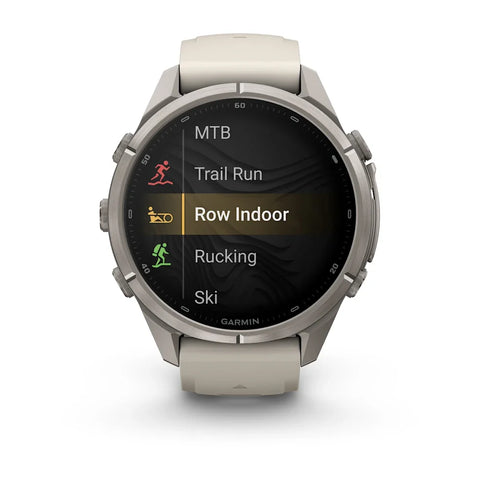A Garmin smartwatch with a round silver-toned metal case and a light beige silicone strap, shown on a clean white background. The watch screen displays a fitness menu with options like MTB, Trail Run, Row Indoor, Rucking, and Ski.