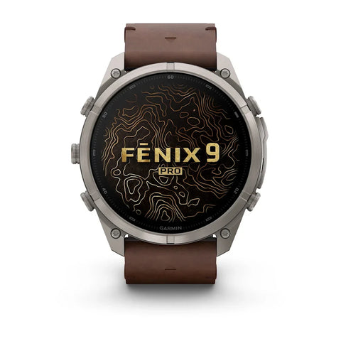 A Garmin Fenix 9 Pro smartwatch with a round silver-toned metal case and a brown leather strap, shown against a plain white background. The watch face displays a black background with gold topographic lines and the text 'FENIX 9 PRO'.