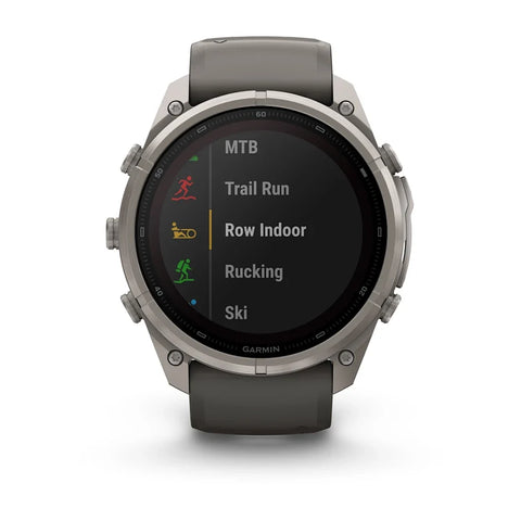A Garmin smartwatch with a round silver-tone metal case and a dark gray silicone strap, shown against a plain white background. The watch face is on and displaying a list of activities including MTB, Trail Run, Row Indoor, Rucking, and Ski.