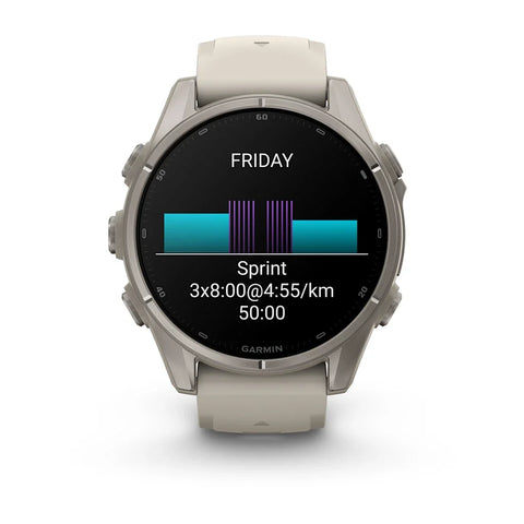 A Garmin smartwatch with a round silver-toned metal case and a light beige silicone strap, shown on a clean white background. The watch face is active with a black display showing a fitness graphic and the word 'FRIDAY'.