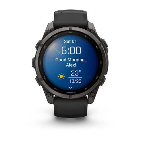 A Garmin smartwatch with a round black and dark gray case and a black strap, shown against a plain white background. The watch face is illuminated with a blue digital display showing the time, weather, and a greeting.