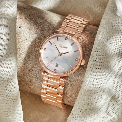 A rose gold-tone women's watch with a round mother-of-pearl dial and matching metal link bracelet, displayed on a textured stone surrounded by draped fabric.