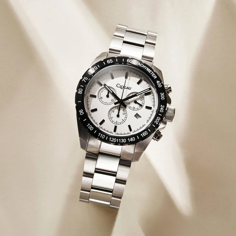 A stainless steel chronograph watch with a white dial, black tachymeter bezel, and matching steel link bracelet, photographed on a warm beige background with soft diagonal shadows.