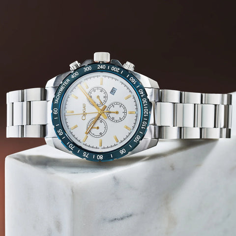 A silver stainless steel wristwatch with a white dial, gold-tone hands and markers, and a dark blue tachymeter bezel, displayed on a white marble block against a dark brown background.