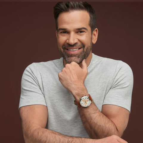 A smiling man with short dark hair and a beard, wearing a light gray textured t-shirt and a brown leather wristwatch with a white dial, posed against a solid dark brown background.