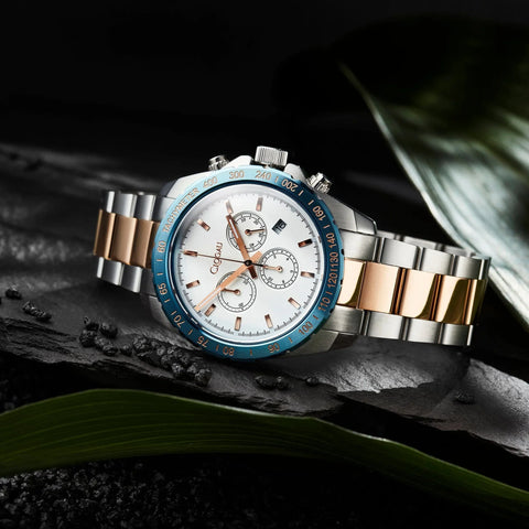 A luxury men's chronograph watch with a two-tone silver and rose gold metal bracelet, a white dial, and a blue bezel, styled on a dark textured surface with green leaves.