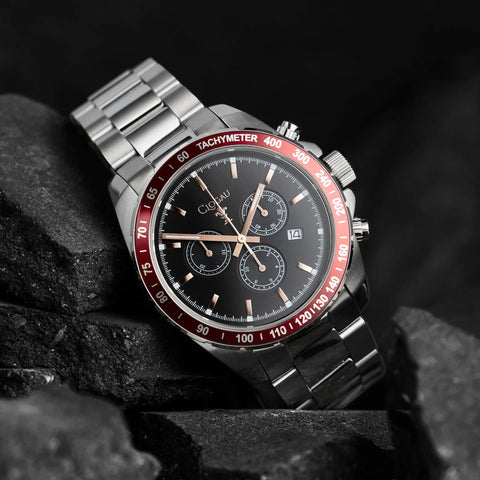 A stainless steel wristwatch with a black dial, red tachymeter bezel, and rose gold-tone hands, shown resting on dark textured rocks.