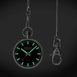 A dark round pocket watch pendant with a black dial and glowing green hour markers, shown hanging from a chain on a dark background. A second detached clasp is displayed to the right.