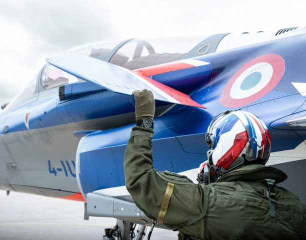 Person in military uniform and helmet standing next to a fighter jet with a French air force emblem.
