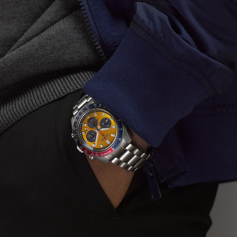 Close-up of a person wearing a silver watch with a yellow face, blue and red accents, on a dark background.