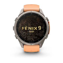 A Garmin Fenix 9 Pro smartwatch with a round silver-toned metal case, a black display screen featuring gold topographic lines and 'FENIX 9 PRO' text, and an orange silicone strap, shown on a clean white background.