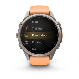 A Garmin smartwatch with a round silver-tone case and an orange strap, shown on a clean white background. The watch screen displays fitness activity options including MTB, Trail Run, Row Indoor, Rucking, and Ski.