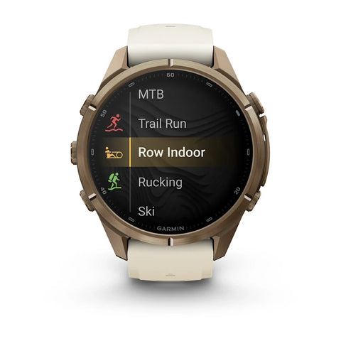 A Garmin smartwatch with a round bronze-toned case and a light beige strap, shown on a clean white background. The watch face displays a fitness menu with options like MTB, Trail Run, Row Indoor, Rucking, and Ski.