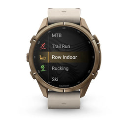 A Garmin smartwatch with a bronze-toned metal case and a light beige leather strap, shown on a clean white background. The watch face displays a fitness menu with options like MTB, Trail Run, Row Indoor, Rucking, and Ski.