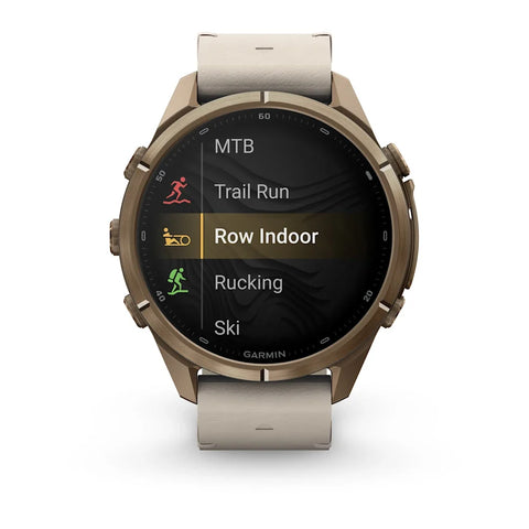 A Garmin smartwatch with a bronze-toned metal case and a light beige leather strap, shown on a clean white background. The watch face displays a fitness menu with options like MTB, Trail Run, Row Indoor, Rucking, and Ski.