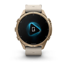 A Garmin smartwatch with a round metallic rose gold case and a light beige silicone strap, shown on a clean white background. The watch face is black with a blue and white digital display.