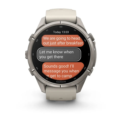 A Garmin smartwatch with a round silver-toned metal case and a light beige silicone strap, shown on a clean white background. The watch face displays a messaging app interface with orange and gray text bubbles.