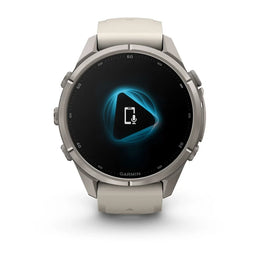 A Garmin smartwatch with a round silver-toned metal case and a light beige silicone strap, shown against a plain white background. The watch face displays a black screen with a glowing blue geometric icon.
