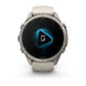 A Garmin smartwatch with a round silver-toned metal case and a light beige silicone strap, shown against a plain white background. The watch face displays a black screen with a glowing blue geometric icon.