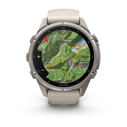A Garmin smartwatch with a round silver-tone metal case and a light beige silicone strap, shown on a clean white background. The watch face is active with a green topographic map display.