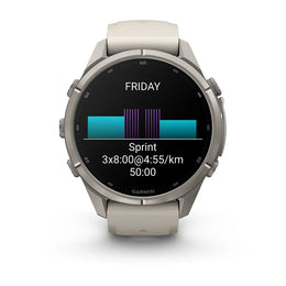 A Garmin smartwatch with a round silver-toned metal case and a light beige silicone strap, shown against a plain white background. The watch face is active with a black display showing a fitness graphic and the word 'FRIDAY'.
