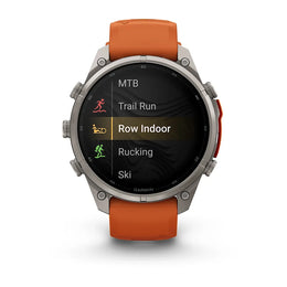 A Garmin smartwatch with a silver-toned metal case and an orange strap, shown on a clean white background. The watch face displays a fitness menu with options like MTB, Trail Run, Row Indoor, Rucking, and Ski.