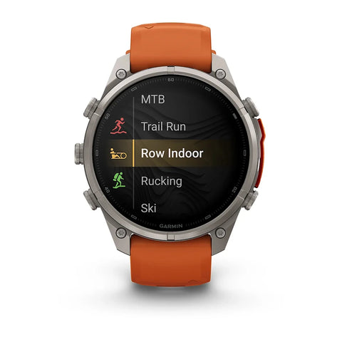 A Garmin smartwatch with a silver-toned metal case and an orange strap, shown on a clean white background. The watch face displays a fitness menu with options like MTB, Trail Run, Row Indoor, Rucking, and Ski.