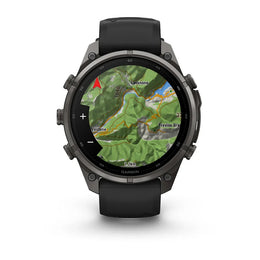 A Garmin smartwatch with a round dark gray metallic case and a black strap, shown on a clean white background. The watch face is active with a green topographic map display.