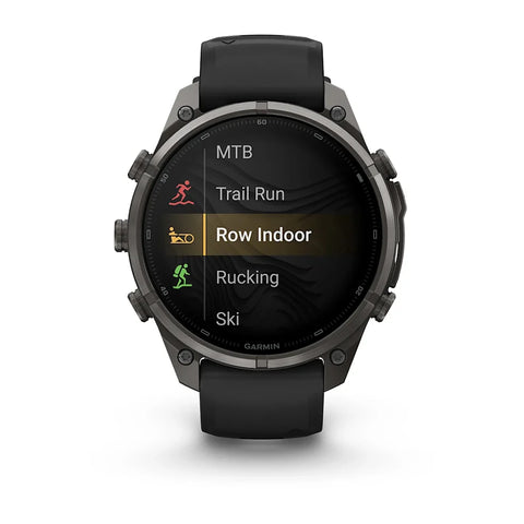 A Garmin smartwatch with a black strap and dark metallic case, shown from the front on a white background. The watch screen displays a fitness menu with options including MTB, Trail Run, Row Indoor, Rucking, and Ski.