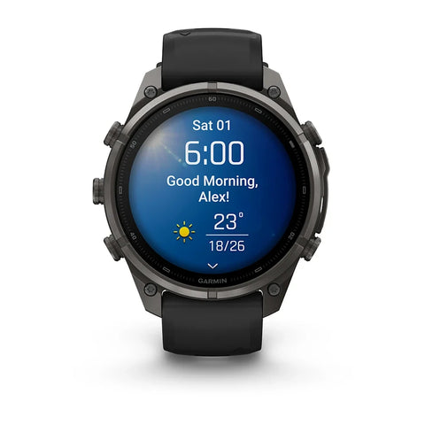 A Garmin smartwatch with a dark gray metallic case and a black strap, shown on a clean white background. The watch face is illuminated with a blue digital display showing the time, weather, and a greeting.
