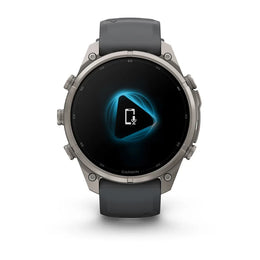 A Garmin smartwatch with a round silver-toned metal case and a dark gray silicone strap, shown against a plain white background. The watch face displays a black screen with a glowing blue geometric icon.