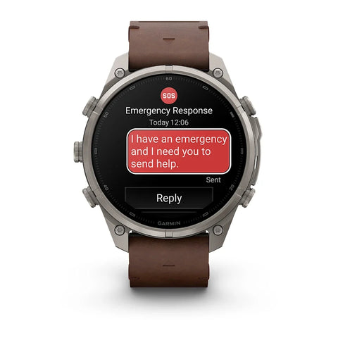 A rugged round Garmin smartwatch with a brown leather strap and a silver-toned metal case, shown on a clean white background. The watch screen displays an emergency message with a red SOS badge and a red text bubble.