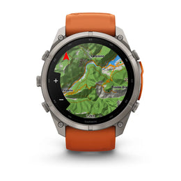 A rugged round smartwatch with a silver-tone metal case and an orange silicone strap, shown front-facing on a white background. The watch face displays a green topographic map with the Garmin logo visible at the bottom.