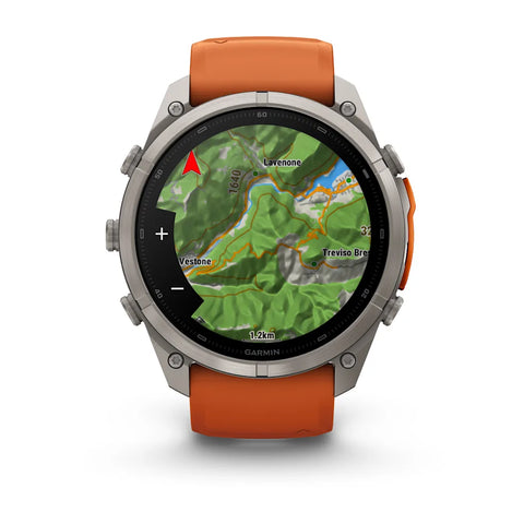 A rugged round smartwatch with a silver-tone metal case and an orange silicone strap, shown front-facing on a white background. The watch face displays a green topographic map with the Garmin logo visible at the bottom.