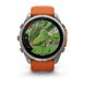 A rugged round smartwatch with a silver-tone metal case and an orange silicone strap, shown front-facing on a white background. The watch face displays a green topographic map with the Garmin logo visible at the bottom.