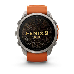 A Garmin Fenix 9 Pro smartwatch with a round silver-toned metal case, a black display screen featuring gold topographic lines and 'FENIX 9 PRO' text, and an orange silicone strap, shown against a plain white background.