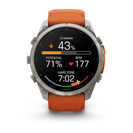A Garmin smartwatch with a silver-toned metal case and an orange silicone strap, shown against a plain white background. The watch face is active with various fitness and heart rate metrics displayed.