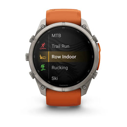 A Garmin smartwatch with a silver-toned metal case and an orange sport band, shown against a plain white background. The watch face displays a fitness menu with options like MTB, Trail Run, Row Indoor, Rucking, and Ski.
