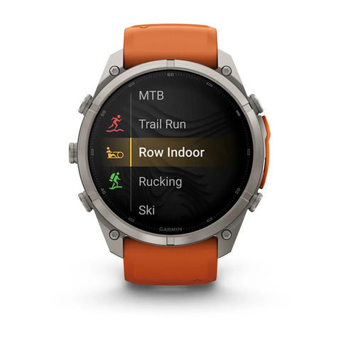 A Garmin smartwatch with a silver-toned metal case and an orange sport band, shown against a plain white background. The watch face displays a fitness menu with options like MTB, Trail Run, Row Indoor, Rucking, and Ski.