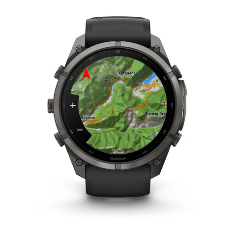 A Garmin smartwatch with a dark gray metallic case and a black strap, shown on a clean white background. The watch face is turned on and displaying a green topographic map with place names like 'Lavigne' and 'Trevio Bridge'.