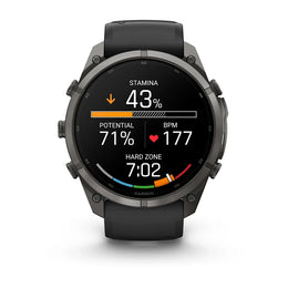 A Garmin smartwatch with a dark gray metallic case and a black strap, shown against a plain white background. The watch face is illuminated with a black background displaying various fitness metrics and colorful graphics.