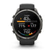 A Garmin smartwatch with a dark gray metallic case and a black strap, shown against a plain white background. The watch face is illuminated with a black background displaying various fitness metrics and colorful graphics.