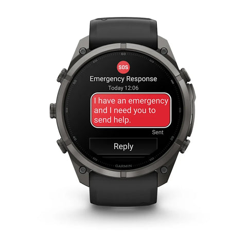 A Garmin smartwatch with a dark gray metallic case and black strap, shown on a white background. The watch screen displays an emergency message with a red SOS icon and a red text bubble reading 'I have an emergency and I need you to send help.