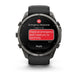 A Garmin smartwatch with a dark gray metallic case and black strap, shown on a white background. The watch screen displays an emergency message with a red SOS icon and a red text bubble reading 'I have an emergency and I need you to send help.