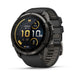 A Garmin Fenix smartwatch with a round black and dark gray case and a textured black strap, shown at a three-quarter angle on a white background. The watch face displays a dark watch UI with yellow accents.