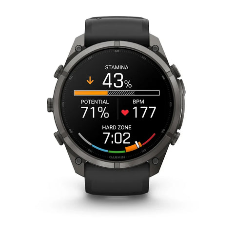 A Garmin smartwatch with a dark gray metallic case and a black strap, shown against a plain white background. The watch face is illuminated with a black background displaying various fitness metrics and colorful graphics.