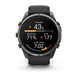 A Garmin smartwatch with a dark gray metallic case and a black strap, shown against a plain white background. The watch face is illuminated with a black background displaying various fitness metrics and colorful graphics.