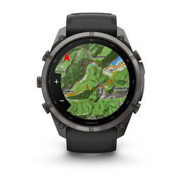 A Garmin smartwatch with a dark gray metallic case and a black strap, shown on a clean white background. The watch face is turned on and displaying a green topographic map with place names like 'Lavigne' and 'Trevio Bridge'.